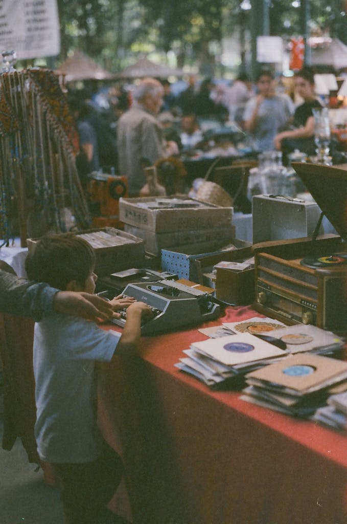 A child curiously explores a vintage stall with gramophones and typewriters at an outdoor market.